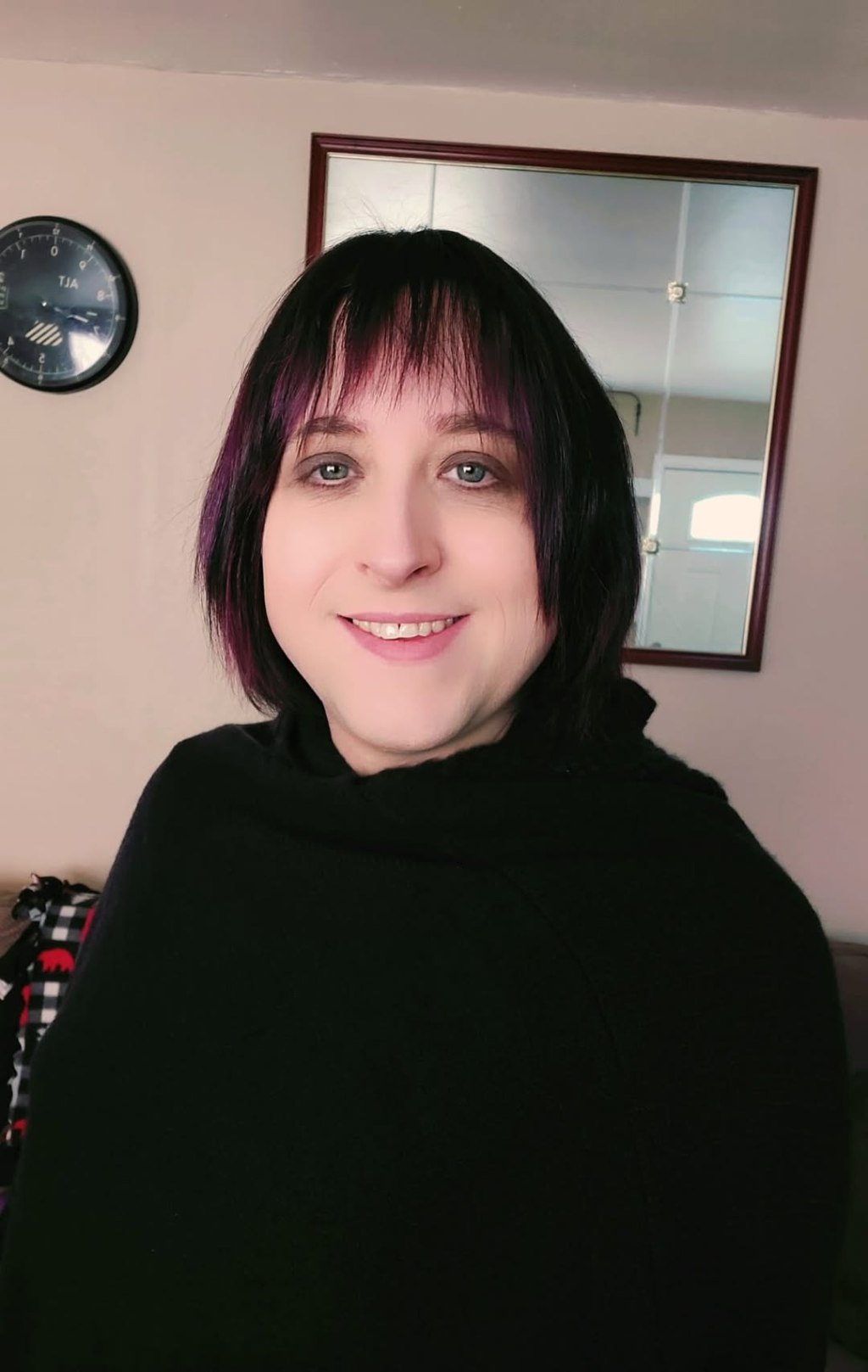 Lily, with shoulder-length dark hair, smiles indoors with a mirror and clock in the background.