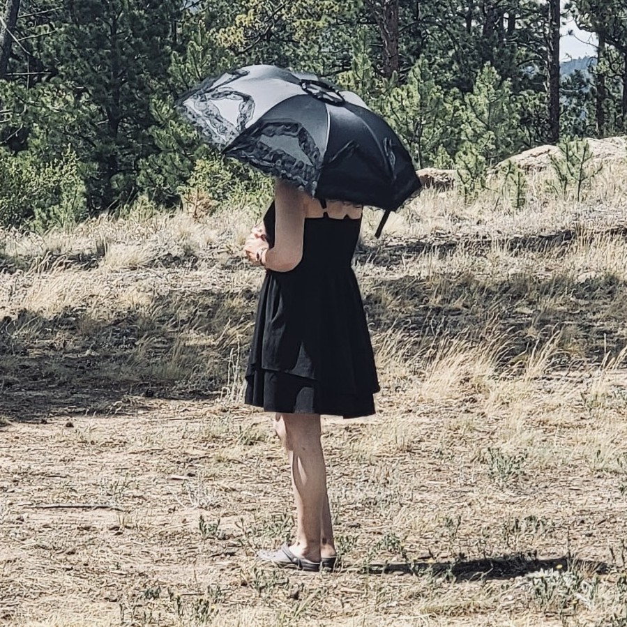 Lily standing alone in a forest, wearing all black and using an umbrella.